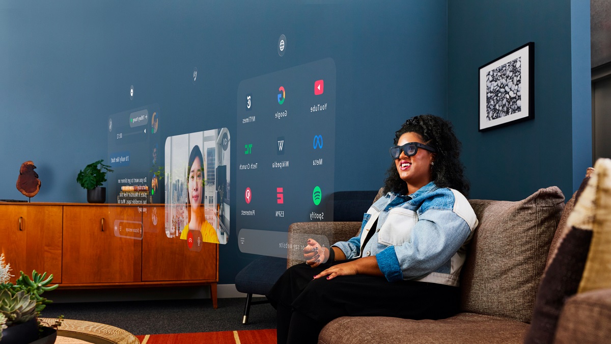 Woman using Meta Orion AR glasses with holographic app interfaces and video call projection in living room.