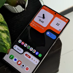 Angled close-up of a Samsung Phone displaying a highly customized dark home screen with split orange widget blocks for the clock and date, arranged next to green plant leaves.